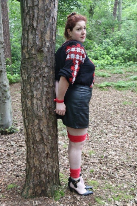 Tjock rödhårig kvinna med munkavle och bunden vid ett träd i en skog